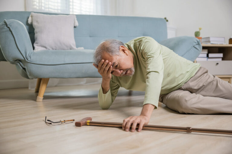 Older man lying on the floor beside a light blue sofa, holding his forehead, with a walking stick and glasses nearby.