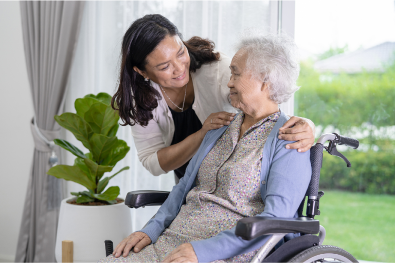 A caregiver talking with an older woman seated in a wheelchair at home.