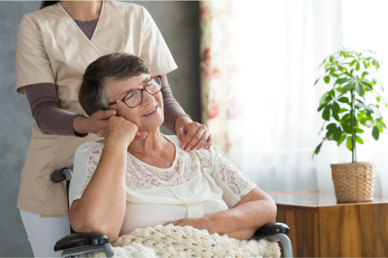 Older woman seated in a wheelchair with a caregiver standing behind her, resting a hand on her shoulder.