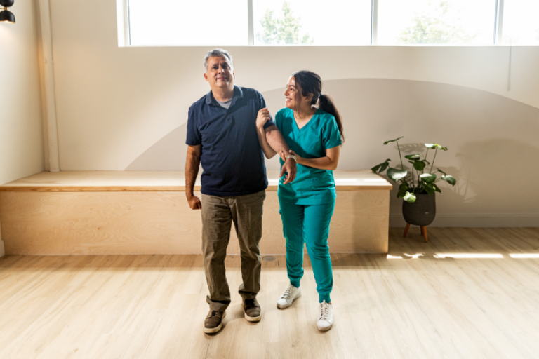 Older man walking indoors while a healthcare worker in green scrubs supports his arm.