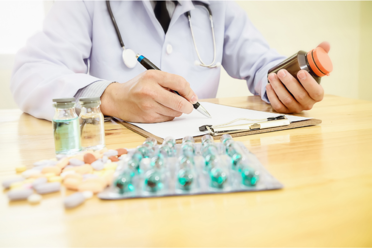 Healthcare professional holding a medicine bottle and writing notes, with tablets and blister packs on the desk.