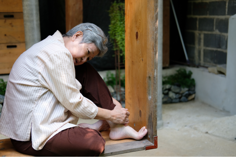 Older woman sitting on a wooden doorstep, holding her ankle with both hands.