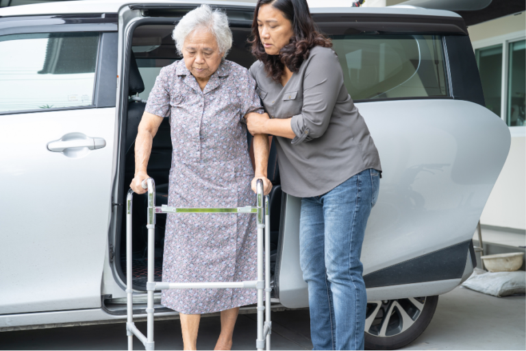 An older woman using a walking frame with support from a caregiver beside a car.