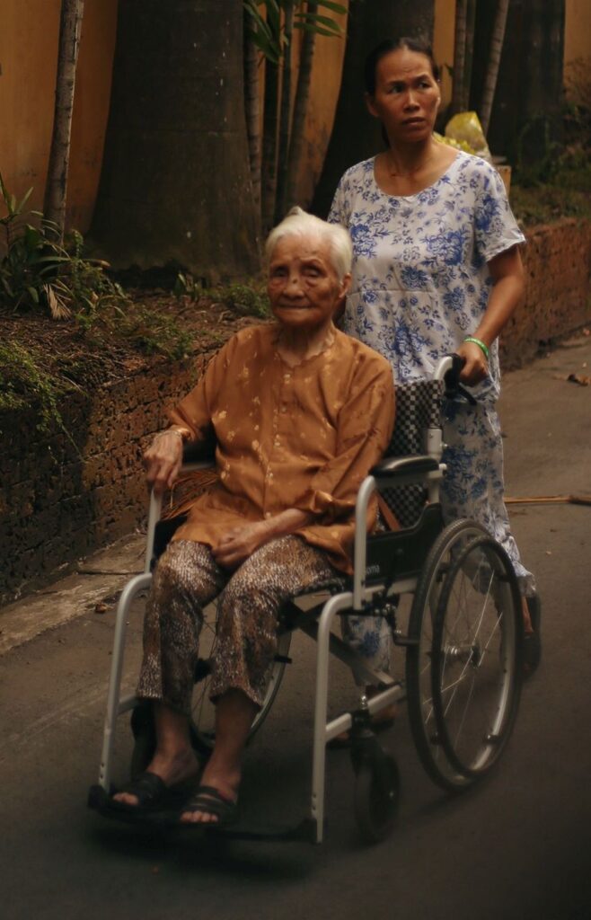 Older woman being pushed in a wheelchair by a companion, illustrating mobility support relevant to geriatric rehabilitation.